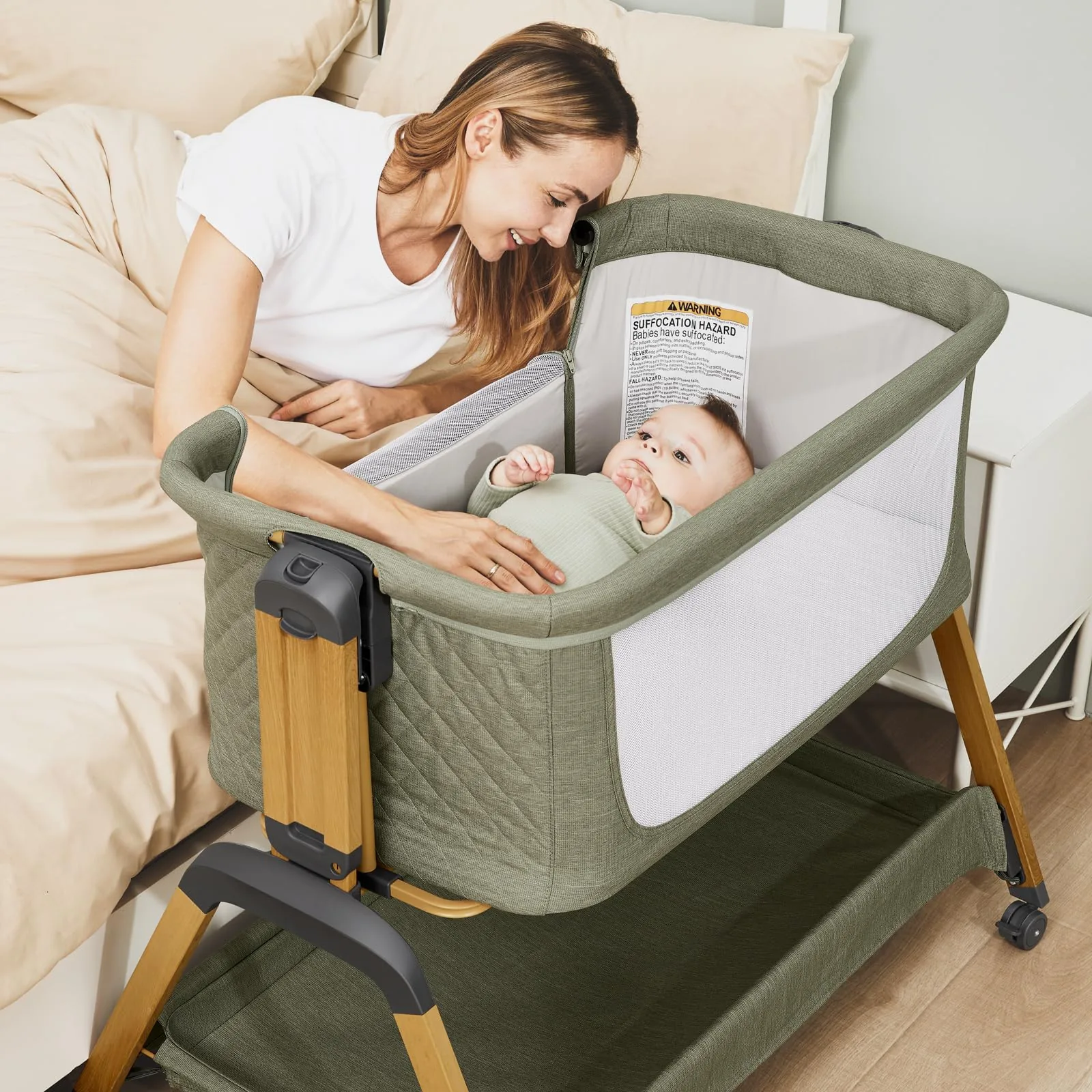 A mother leaning over a sage green Jimglo bassinet placed against her bed, smiling at the baby lying inside